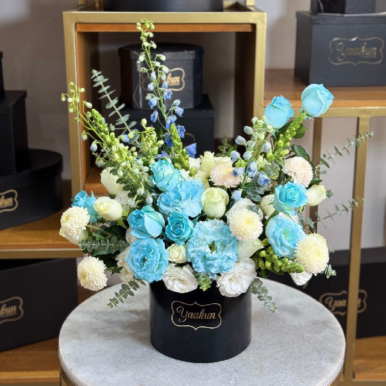 Caja circular negra con 25 rosas blancas, lisianthus y delfinio azul